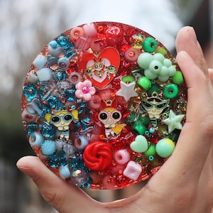 Puede incluir: Una pieza de arte redonda, roja, de resina con un diseño en forma de corazón que presenta a las Chicas Superpoderosas. La pieza está llena de cuentas de colores, purpurina y otros adornos.