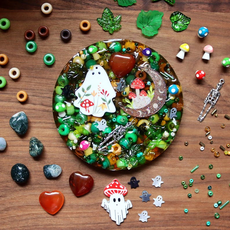 May include: A whimsical resin coaster with a variety of decorative elements. The coaster features a central design with a ghost, mushrooms, and a crescent moon. Surrounding the coaster are beads, stones, and other small decorative items. The color palette includes green, brown, red, and white.