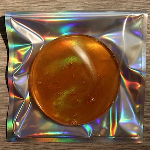 May include: A round, amber-coloured product is presented within a holographic, iridescent package. The product has a glossy, gel-like appearance. The package is sealed and reflects a spectrum of colours.