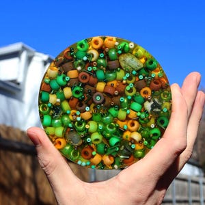 May include: A round, colorful coaster with a mosaic design. The coaster is filled with a variety of beads in shades of green, brown, yellow, and clear. The coaster is held in a hand against a blue sky background.