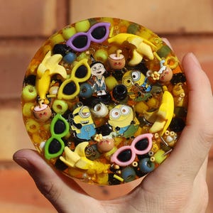 Può includere: Un sottobicchiere in resina gialla con una varietà di ciondoli colorati, tra cui personaggi dei cartoni animati, occhiali da sole, banane e perline.