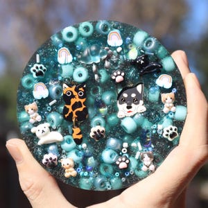 May include: A round, teal-colored resin art piece filled with small beads and various animal-themed charms. The charms include cats, dogs, paw prints, and rainbows. The piece is held in a person's hand.