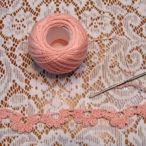 May include: Pink crochet thread wound around a spool, a crochet hook, and a crocheted lace trim with a scalloped edge.