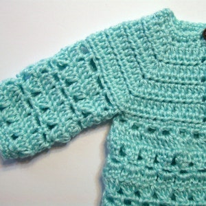 May include: A light blue crocheted cardigan with a button closure. The cardigan has a simple stitch pattern and is made with a soft yarn.