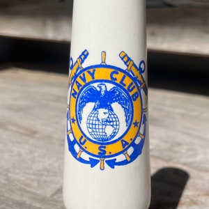 May include: White ceramic vase with a blue and yellow Navy Club logo featuring an eagle, globe, and anchors. The vase has a flared base.
