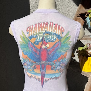 May include: A sleeveless, light purple t-shirt featuring a colorful parrot design with the words "Hawaiian Tropic". The parrot has red, blue, and green feathers, with a tropical beach scene in the background.