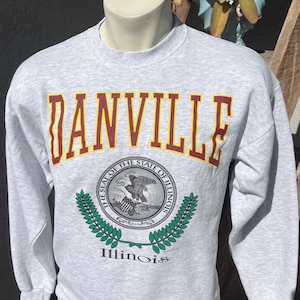 Puede incluir: Sudadera gris con un gráfico "DANVILLE" en granate y amarillo y un sello del estado de Illinois con una corona de laurel verde y la palabra "Illinois" debajo.