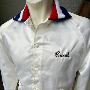 May include: White satin jacket with a red, white, and blue striped collar. The jacket has a button-up front, two pockets, and the name "Carol" embroidered in black script on the chest. The jacket is displayed on a mannequin.