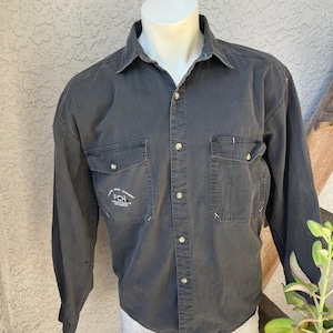 Puede incluir: Una camisa gris oscuro de manga larga con dos bolsillos en el pecho y botones. La camisa tiene cuello y el texto "PCH" es visible en el bolsillo izquierdo. La camisa se exhibe en un maniquí blanco.