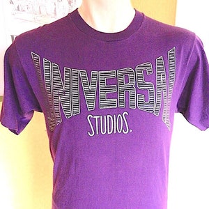 May include: Purple t-shirt with a white "UNIVERSAL STUDIOS." graphic printed on the front.