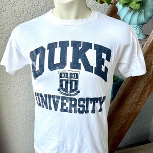 Peut inclure: T-shirt blanc avec le mot "DUKE" en lettres majuscules bleu marine sur la poitrine. En dessous, un écusson bleu marine et le mot "UNIVERSITY". Le t-shirt est présenté sur un mannequin.