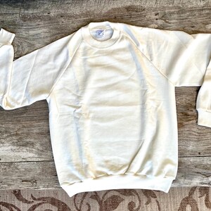 May include: Cream-colored sweatshirt with long sleeves and a crew neck. The raglan sleeves extend from the collar to the underarm. The cuffs and hem are ribbed. The sweatshirt is made of a soft, comfortable material.