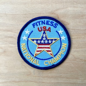May include: A blue and gold embroidered patch with a star and the words "Fitness USA National Champion".