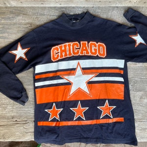 May include: A black long-sleeve shirt with a white and orange striped design. The shirt features a large white star and the word "CHICAGO" in white letters. There are also smaller white stars on the sleeves and bottom of the shirt.