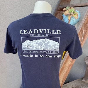 Könnte beinhalten: Marineblaues T-Shirt mit einem Aufdruck auf der Rückseite mit der Aufschrift "LEADVILLE COLORADO" über einer Bergillustration. Der Text "Mt. Elbert elev. 4.399 m" ist ebenfalls enthalten. Das Shirt sagt auch "I made it to the top!"