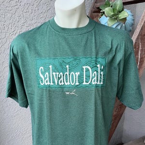 May include: Green striped t-shirt with the name "Salvador Dali" in white block letters on a rectangular teal patch. The shirt has short sleeves and a crew neck. A signature is below the name.