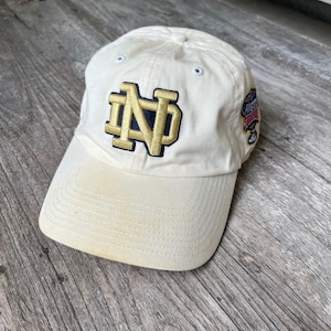 Op de afbeelding: Crèmekleurige baseballpet met de letters "ND" in goud en marineblauw. De pet heeft een gebogen klep en een kleine patch aan de zijkant. De hoed is gemaakt van een zacht materiaal, ideaal voor casual wear.