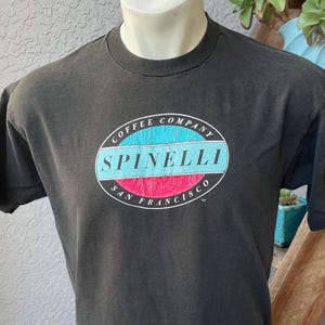 Può includere: Maglietta nera con un logo circolare che recita "Coffee Company Spinelli San Francisco". Il logo ha una combinazione di colori blu, rosa e bianco.
