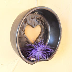 May include: A dark grey ceramic wall planter with a heart-shaped cut-out and a purple air plant. The planter has a rounded shape and is mounted on a light yellow wall. The heart cut-out allows light to pass through.