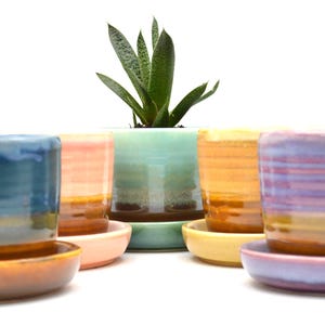 May include: Five colorful ceramic planters with matching saucers. The planters are striped with shades of blue, green, pink, orange, and purple. The saucers are the same colors as the planters.