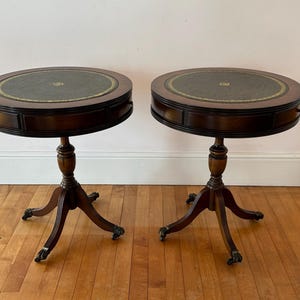Puede incluir: Dos mesas de tambor antiguas con marcos de madera oscura. Cada mesa tiene una tapa redonda con una inserción de cuero verde y un borde dorado. Las mesas se apoyan en patas de trípode con pequeñas ruedas.