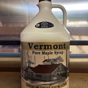 May include: A white plastic gallon jug of Vermont Pure Maple Syrup with a brown cap and a label featuring a cartoon illustration of a maple syrup farm. The label also includes the text "Vermont Pure Maple Syrup" and "1 GAL 128 FL OZ 3.78 L".