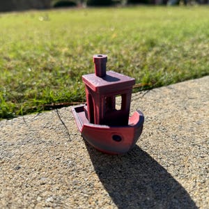 Könnte beinhalten: Ein kleines, 3D-gedrucktes Spielzeugboot in Rot- und Lilatönen. Das Boot hat eine Kabine mit Schornstein und einen abgerundeten Bug. Das Spielzeug liegt auf einer Betonoberfläche, mit Gras im Hintergrund.