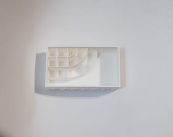 May include: A white plastic organizer with multiple compartments. The organizer has a rectangular shape with a curved section and a larger open area. The compartments are various sizes, suitable for storing small items.