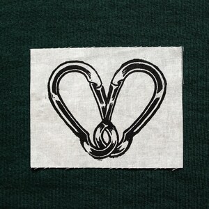 May include: A black and white graphic of two carabiners forming a heart shape, printed on a square of light gray fabric. The fabric is set against a dark green background.