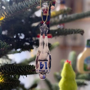 May include: A clear acrylic Christmas ornament featuring a football player in a white uniform with the number 14 and the name "ST BROWN". The ornament hangs upside down from a gold chain on a Christmas tree.