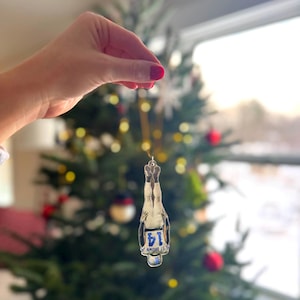 May include: Clear glass Christmas ornament shaped like a champagne bottle. The ornament has a silver metal top and a blue and white label that reads "14 ST BROWN". The ornament is hanging in front of a decorated Christmas tree.