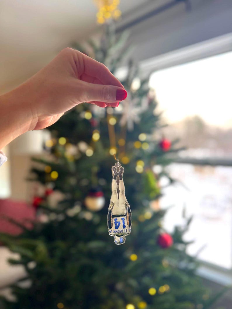 May include: Clear glass Christmas ornament shaped like a champagne bottle. The ornament has a silver metal top and a blue and white label that reads "14 ST BROWN". The ornament is hanging in front of a decorated Christmas tree.