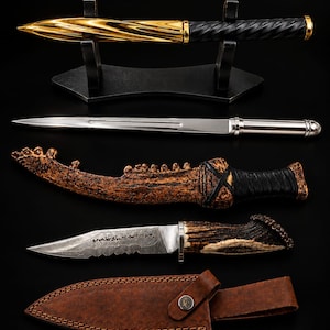 Könnte beinhalten: Eine Sammlung von Gegenständen, darunter ein gold-schwarzer gedrehter Stift auf einem Ständer, ein silberner Brieföffner, eine strukturierte braune Scheide mit schwarzem Griff, ein Messer mit Wellenschliff und Hirschhorngriff sowie eine braune Lederscheide mit Logo.