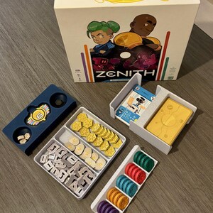 Puede incluir: Un juego de mesa titulado Zenith que incluye una caja con personajes ilustrados, una bandeja azul oscuro con una ficha central, una bandeja blanca con fichas amarillas y grises, un tarjetero blanco y una bandeja con pilas de discos de colores.
