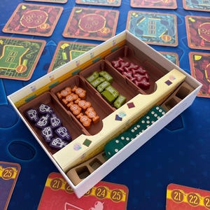 Puede incluir: Un juego de mesa con cartas, dados y piezas de juego. Las piezas del juego están en una caja blanca con compartimentos marrones. Las cartas son rojas y verdes con varios diseños y números. Los dados son verdes con puntos blancos.