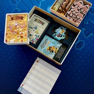 Op de afbeelding: Een bordspel met kaarten, fiches en een scoreblad. De open speldoos toont kaarten met illustraties van kastelen en dorpen. Fiches en spelstukken zijn in compartimenten gerangschikt. De achtergrond is een blauw oppervlak met een dobbelsteenpatroon.