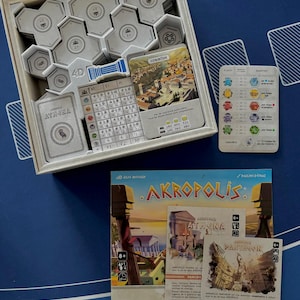 Peut inclure: Jeu de société "Akropolis" avec des pièces, des cartes et une boîte. La boîte contient des tuiles hexagonales grises et des cartes avec des illustrations et du texte. Les cartes de jeu comportent les mots "ATHENA" et "CORINTHE".