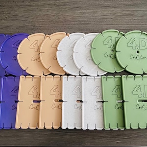 Può includere: Una collezione di rocchetti per filati circolari e rettangolari in viola, beige, bianco e verde salvia. Ogni pezzo presenta il testo in rilievo 4D Création, un foro centrale e bordi dentellati per fissare il filo, disposti in due file.