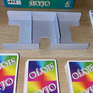 Puede incluir: Un juego de mesa con un tarjetero azul claro, cartas y una caja. Las cartas tienen un diseño de arcoíris y la palabra "SKYJO" impresa. El tarjetero tiene tres compartimentos.