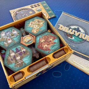 Peut inclure: Une boîte de jeu de société contenant des tuiles hexagonales avec des paysages illustrés, un jeton circulaire et des pièces cylindriques marron. Un livret de règles intitulé Diluvium et un plateau de jeu sont posés près de la boîte jaune.