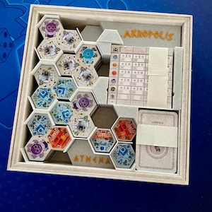 Peut inclure: Une boîte de jeu de société carrée contenant des tuiles hexagonales avec des illustrations architecturales colorées, un carnet de score avec le texte AKROPOLIS et un jeu de cartes. L'intérieur présente des compartiments moulés avec le mot ATHENA.