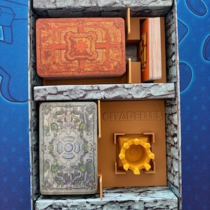 Puede incluir: Una caja de cartón marrón con un patrón de piedra en el exterior. La caja está abierta y contiene un inserto de juego con compartimentos para cartas y una torre. El inserto está etiquetado "Citadelles".