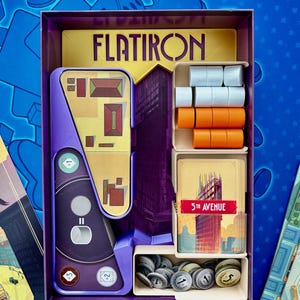 Pode incluir: Uma caixa de jogo de tabuleiro intitulada "FLATIRON" com vários componentes. A caixa contém cartas com a etiqueta "5th Avenue", cilindros de madeira em azul e laranja e moedas de metal. O tabuleiro do jogo apresenta um design de elevador roxo.
