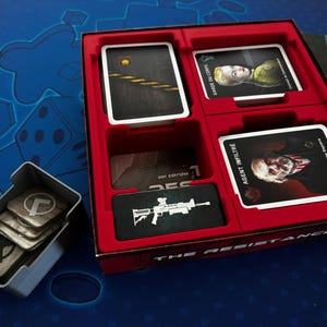 Op de afbeelding: Een rode speldoos met compartimenten voor kaarten en spelstukken. De kaarten bevatten afbeeldingen en tekst, waaronder "AGENT INFILTRÉ" en een geweer. Een aparte grijze container bevat extra spelstukken. Het spel heet "THE RESISTANCE."
