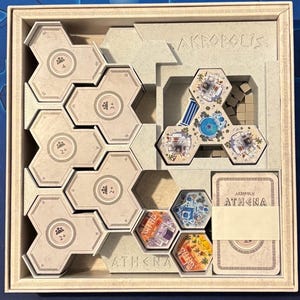 Peut inclure: Une boîte de jeu beige avec un insert blanc pour ranger les pièces du jeu. L'insert comporte une section intitulée "Athena" et une section intitulée "Akropolis". La boîte contient un plateau de jeu, des cartes et d'autres pièces de jeu.