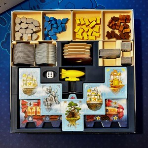 Peut inclure: Un jeu de société avec divers composants : jetons en bois, cartes illustrées, un dirigeable jaune et un dé. Les pièces sont rangées dans une boîte noire avec des compartiments beiges, sur un fond bleu. Jeu de société.