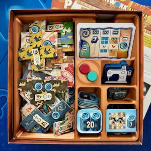 Op de afbeelding: Een Patchwork-spelbord met een verscheidenheid aan kleurrijke stoffen, knopen en score-tokens. Het bord heeft een houten frame en is verdeeld in compartimenten voor verschillende spelonderdelen. Het spel is ontworpen voor 2 spelers en is een populaire keuze voor families en vrienden.