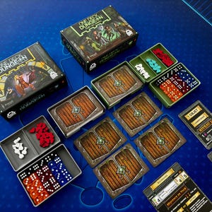 Może przedstawiać: Zestaw gier planszowych "One Deck Dungeon" zawierający pudełka do gry, kości, karty i żetony. Elementy gry są ułożone na niebieskiej powierzchni. Pudełka do gry zawierają ilustracje postaci fantasy i tytuł gry.