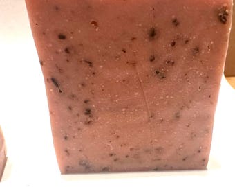 May include: A rectangular bar of handmade soap in a soft pink hue, speckled with dark flecks. The soap has a slightly irregular shape, suggesting a natural, artisanal quality. The texture appears smooth with visible inclusions.