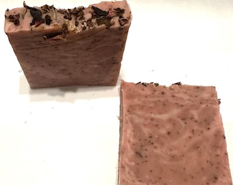 May include: Two bars of handmade soap. The soap is a dusty rose color with a marbled texture. The top of the soap is decorated with dried flowers and herbs. The soap is rectangular in shape.